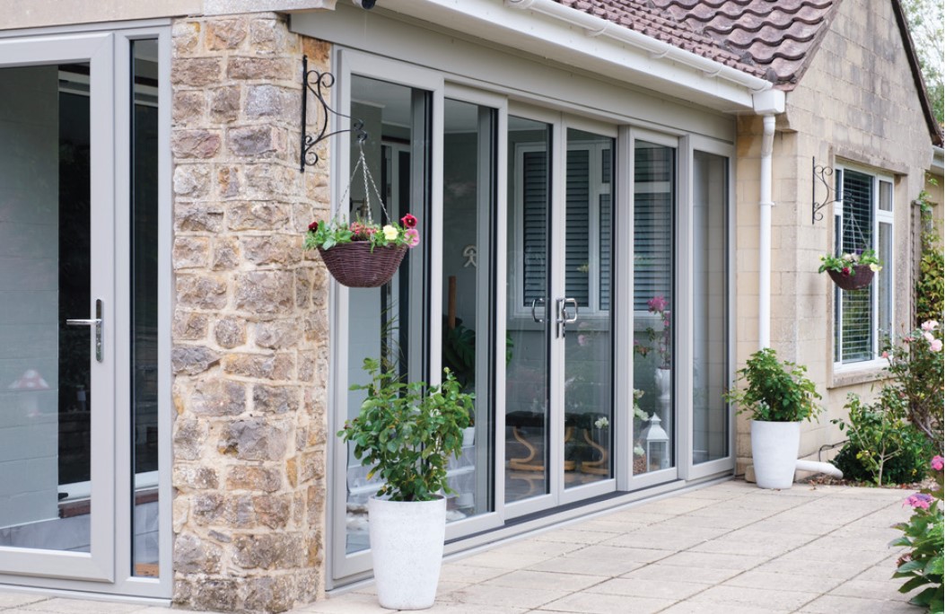 Exterior view of a residential home featuring large light grey triple track sliding patio glass doors installed along a stone and brick patio area with potted plants.